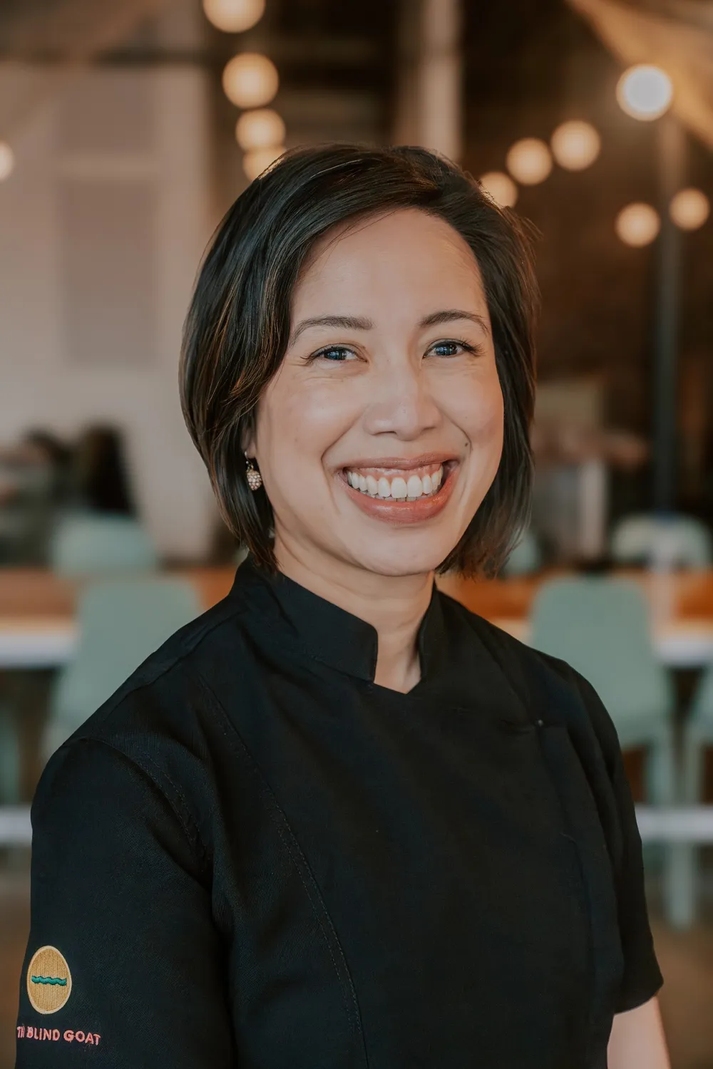 Christine Ha smiling in a black chef’s coat with The Blind Goat logo on the sleeve.